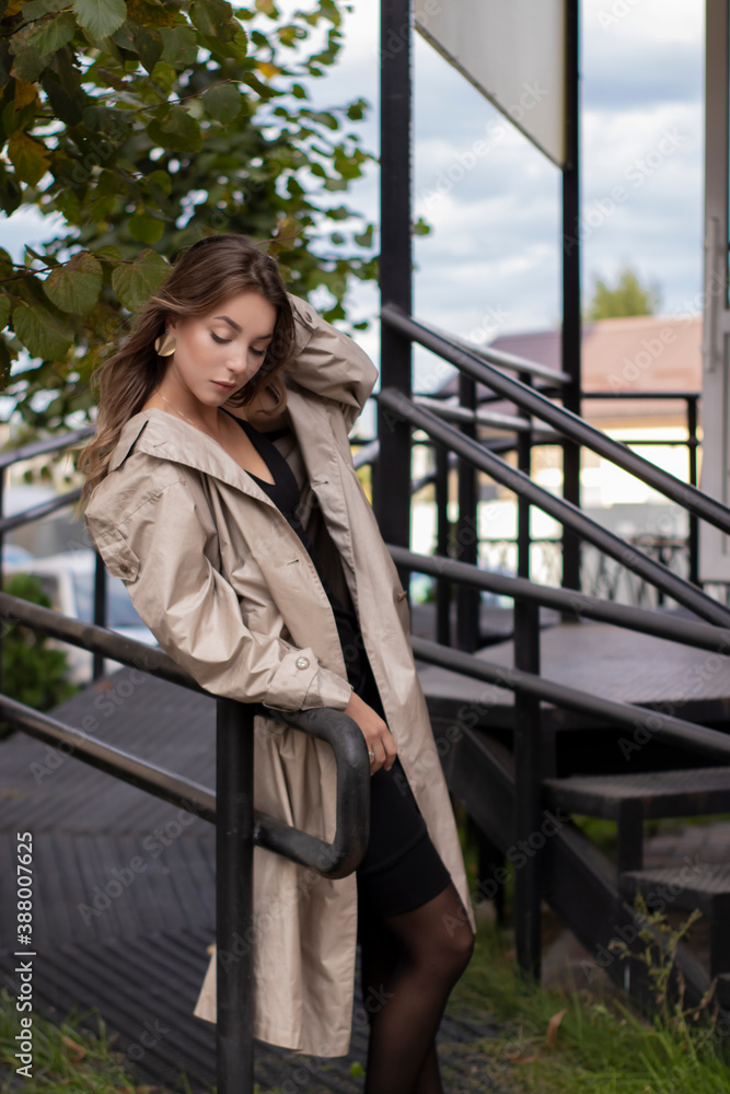 beautiful slim young brunette in fashionable trench coat and black dress walking near stairs. autumn, yellow leaves. . beautiful slim young brunette in fashionable trench coat and black dress walking near stairs. autumn, yellow leaves. .