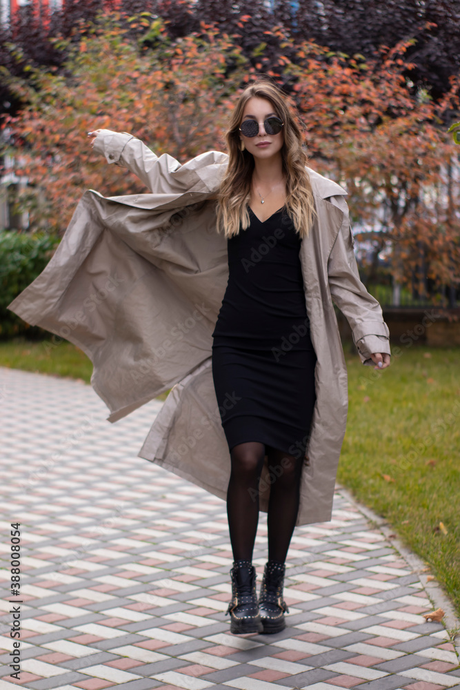 attractive slim young brunette in fashionable trench, black dress and sunglasses in park. autumn, yellow leaves.