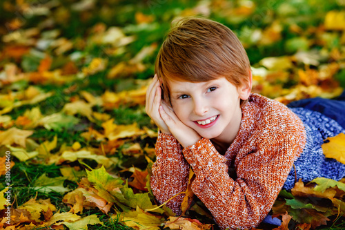 a small red haired boy in a sweater looks at the camera and smiles lying in the fallen yellow leaves of an autumn Park