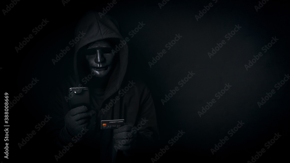 Dangerous anonymous hacker man in hooded and mask use smartphone and credit card, break security data and hack password with personal data Bank account. Internet crime, cyber attack security concept