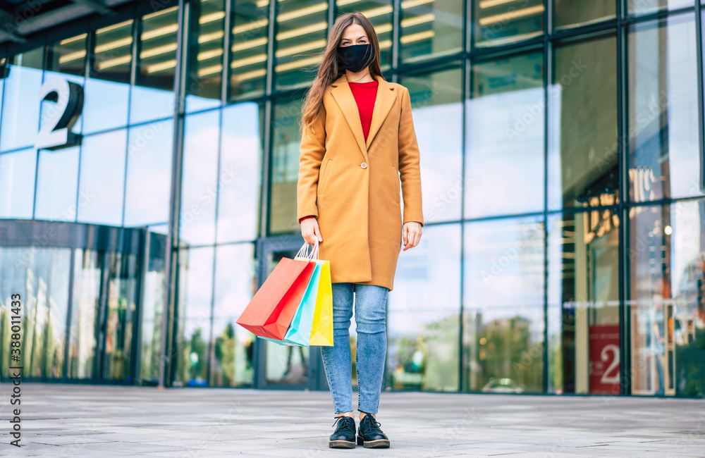Full length photo of young confident woman in coat and safety medical mask with shopping bags is posing on the mall background