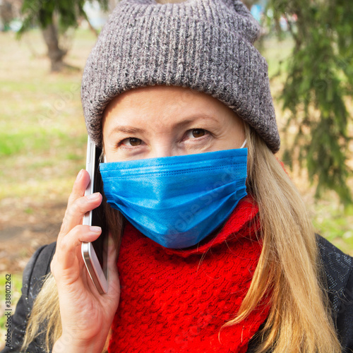 A woman in a knitted hat and a medical mask speaks on a cell phone in the park. Coronavirus concept. Social distance © Svetlana Khor