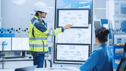 On a Meeting Chief Industrial Engineer Wearing Safety Jacket, and Hardhat Reports to a Group of Specialists, Managers, Uses Interactive Digital Whiteboard to Show New Eco Friendly Engine Technology