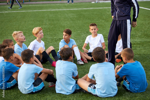 friendly kid boys have rest with trainer during football competition, confident male trainer give advice, explain and have talk with sportive boys, in stadium