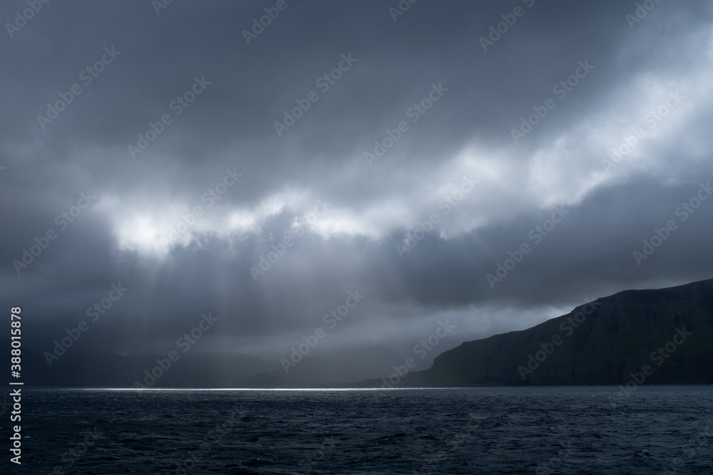 Sailing by Suðuroy island in dramatic light. Moody clouds on sky and sun rays (god rays) coming through and lighting up the ocean. Faroe Islands.