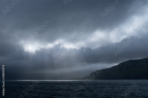 Sailing by Suðuroy island in dramatic light. Moody clouds on sky and sun rays (god rays) coming through and lighting up the ocean. Faroe Islands.