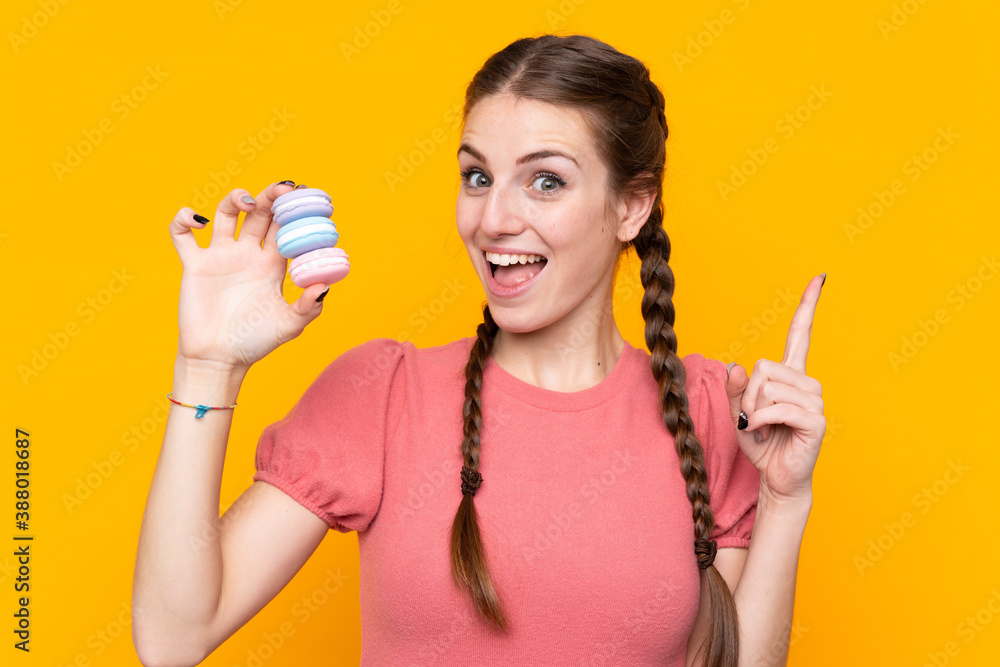 Young woman over isolated yellow background holding colorful French macarons and pointing up a great idea Young woman over isolated yellow background holding colorful French macarons and pointing up a great idea