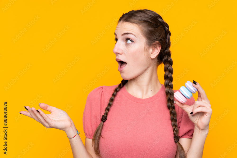 Young woman over isolated yellow background holding colorful French macarons and with surprise expression Young woman over isolated yellow background holding colorful French macarons and with surprise expression