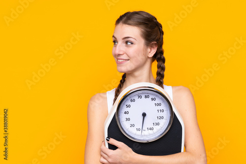 Young woman over isolated yellow background with weighing machine and looking side