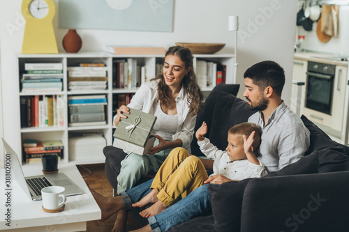 young happy family with one kid sitting on sofa and opening gifts while having video call on computer