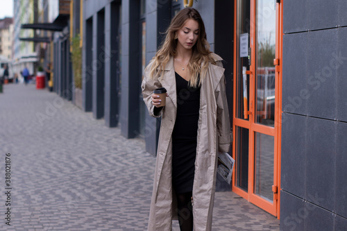 beautiful slim young brunette in fashionable trench coat and black dress walking through the street. autumn, yellow leaves