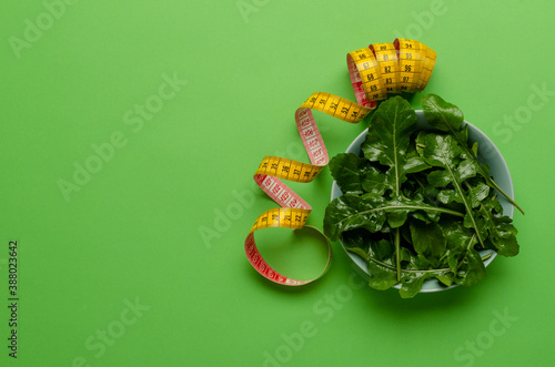 Top view of blue bowl full of juicy fresh arugula leaves and yellow measuring tape on the green surface.Empty space.Concept of healthy eating and diet
