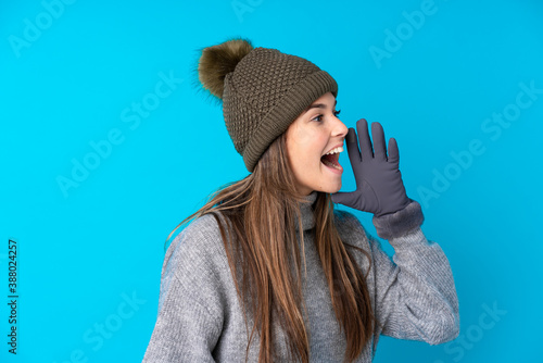 Teenager girl with winter hat over isolated blue background shouting with mouth wide open