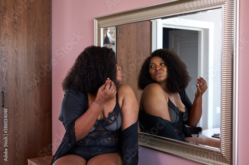 beautiful overweight african lady looks at mirror in living room, loves herself, enjoy being fat. sits on table in lingerie photo