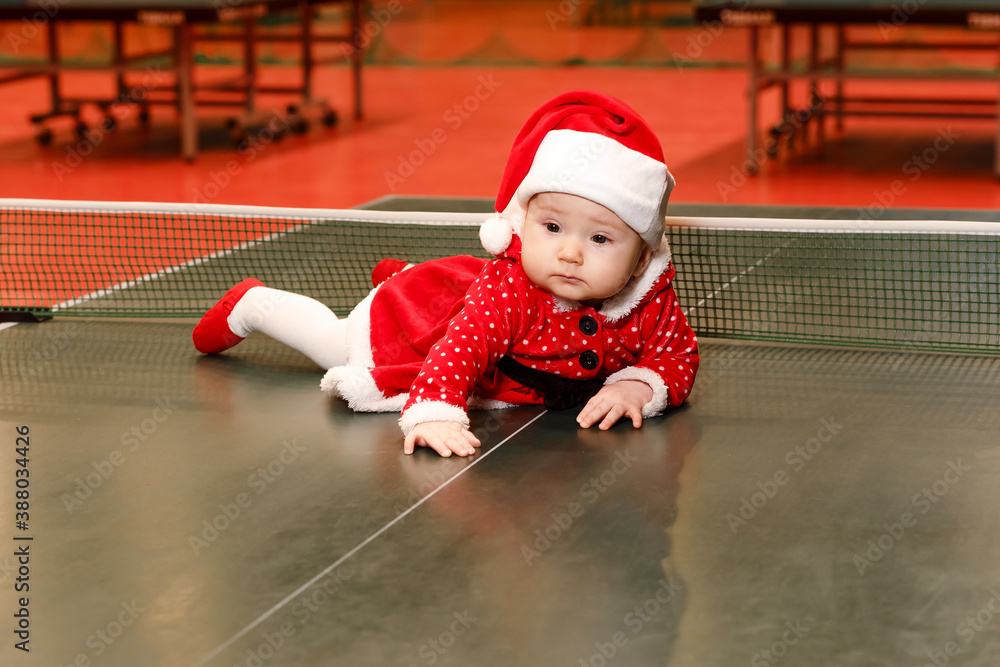 Φωτογραφία six-month-old baby girl in a red Santa suit lies on her stomach on a tennis tabl