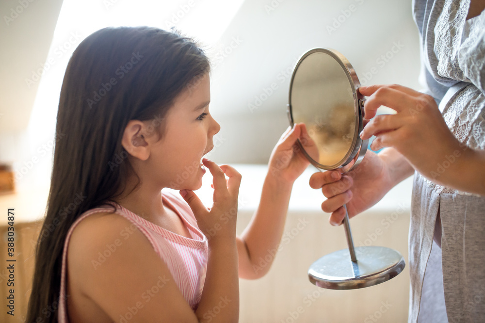 Φωτογραφία Small girl with mother in bathroom indoors, looking in mirror at baby loose tooth