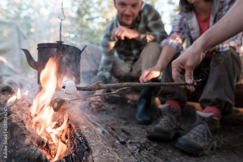 Group of friends fry sweet marshmallows over a fire, over the flame of which a kettle hangs, in a camping camp, in the forest.