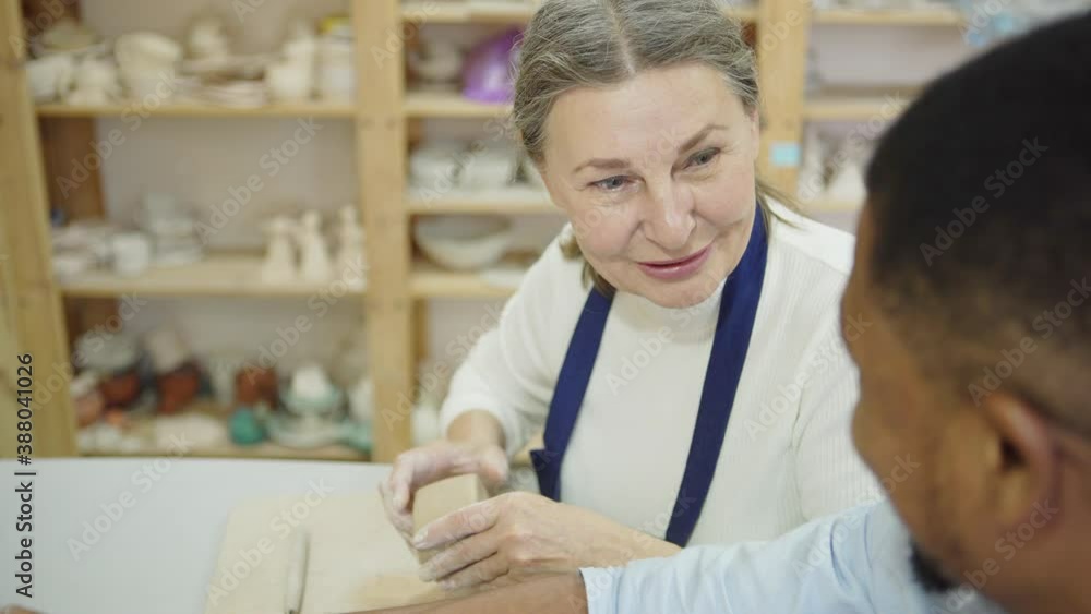 Tilt up side view of senior female ceramic artist teaching young Black man how to shape clay during workshop in pottery studio Tilt up side view of senior female ceramic artist teaching young Black man how to shape clay during workshop in pottery studio