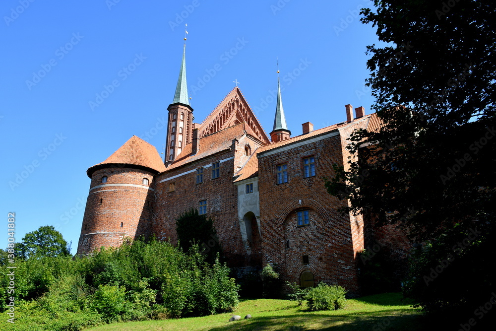 Fototapeta premium View of an old medieval castle made out of red brick with many towers, walls, and the top being a remnant of a church seen next to a public park on a cloudless summer day on a Polish countryside