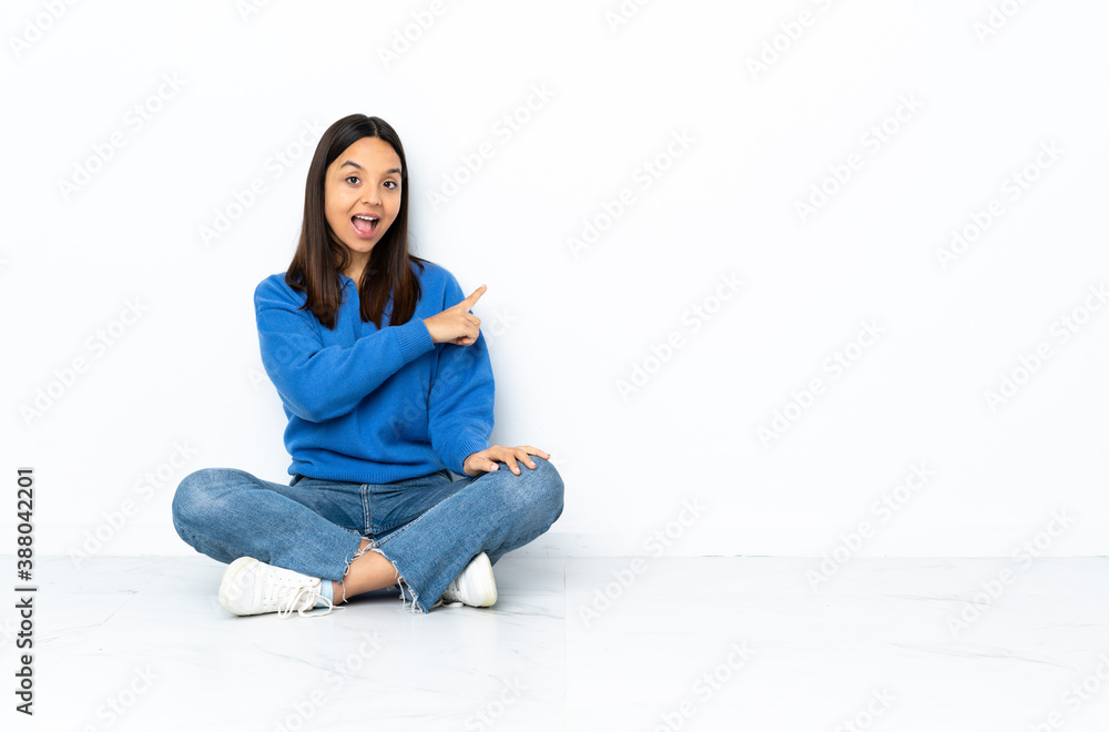 Young mixed race woman sitting on the floor isolated on white background surprised and pointing side Young mixed race woman sitting on the floor isolated on white background surprised and pointing side