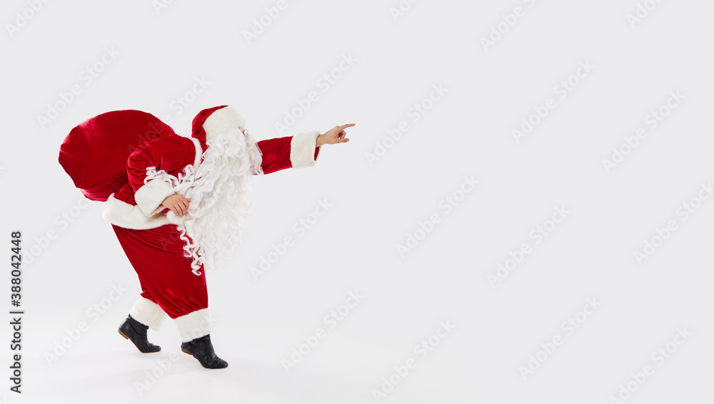 Joyful Santa walks widely with a bag of gifts on his back on a white background. Shows his hand to the right to an empty space for text. New Year and Christmas holidays and sales, banner Joyful Santa walks widely with a bag of gifts on his back on a white background. Shows his hand to the right to an empty space for text. New Year and Christmas holidays and sales, banner