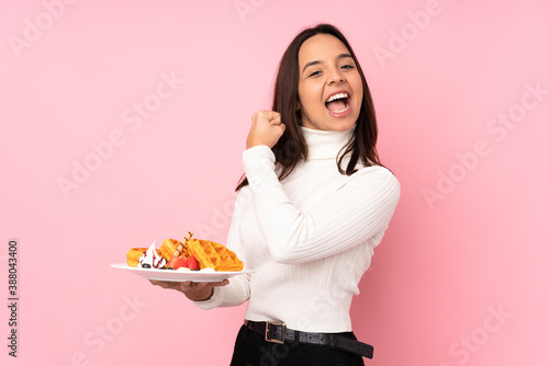 Obraz na plátne Young brunette woman holding waffles over isolated pink background celebrating a
