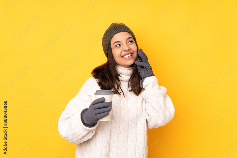 Young woman with winter hat over isolated yellow background holding coffee to take away and a mobile Young woman with winter hat over isolated yellow background holding coffee to take away and a mobile