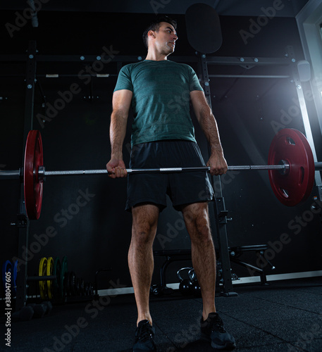 Tall dark man with green t shirt lifting barbell with red weights on a dark gym.