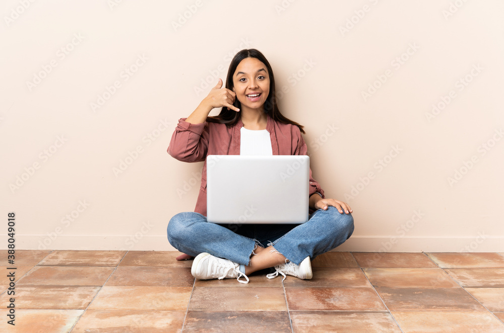 Young mixed race woman with a laptop sitting on the floor making phone gesture. Call me back sign Young mixed race woman with a laptop sitting on the floor making phone gesture. Call me back sign