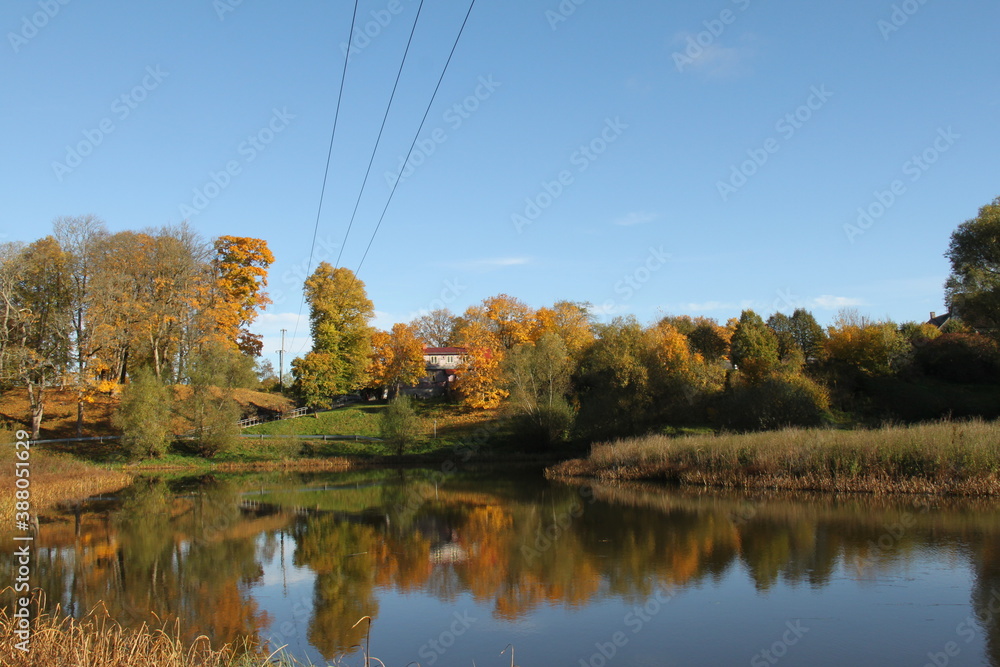 Power lines above the pond in autumn, trees and blue sky mirroring, public park near church in Aizpute, Latvia, Europe Power lines above the pond in autumn, trees and blue sky mirroring, public park near church in Aizpute, Latvia, Europe