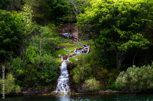 A man-made waterfall flows into the St. Lawrence River from the island of Boldt Castle in the Thousand  1000  Islands  Ontario  surrounded by lush green nature  as seen from a boat tour.