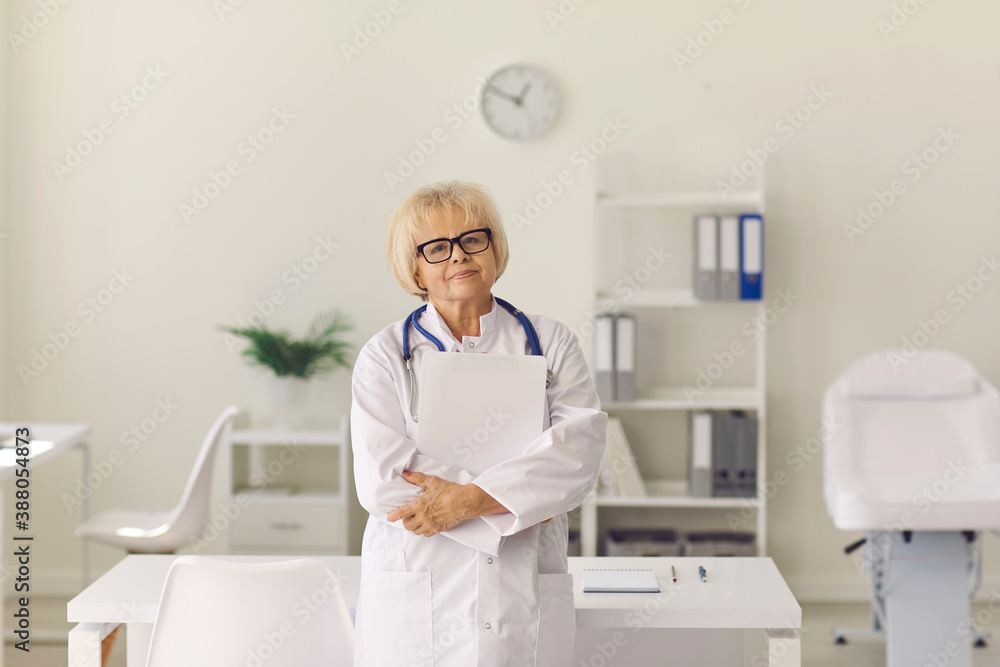 Portrait of a senior female doctor with documents in her hands standing in a modern hospital office.