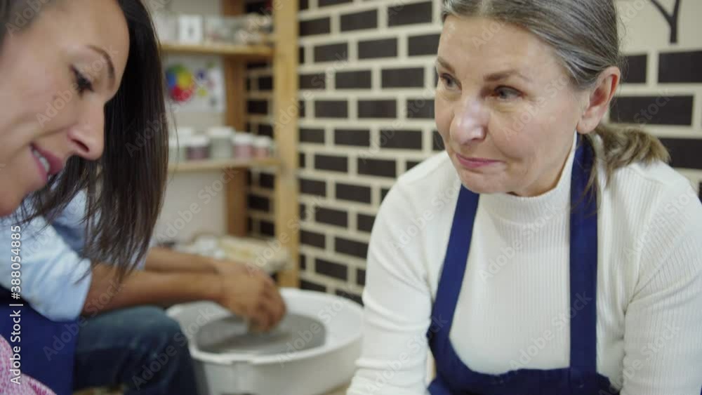 Tilt up and down shot of senior female pottery artist helping middle aged student to throw pot on pottery wheel in ceramic art studio Tilt up and down shot of senior female pottery artist helping middle aged student to throw pot on pottery wheel in ceramic art studio