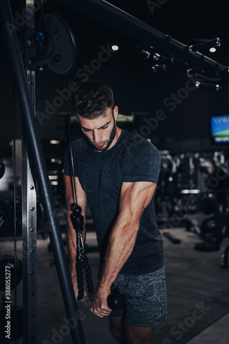 Good looking handsome male athlete exercising in modern fitness gym. Dark muddy light with strong shadows. Indoors sport concept.