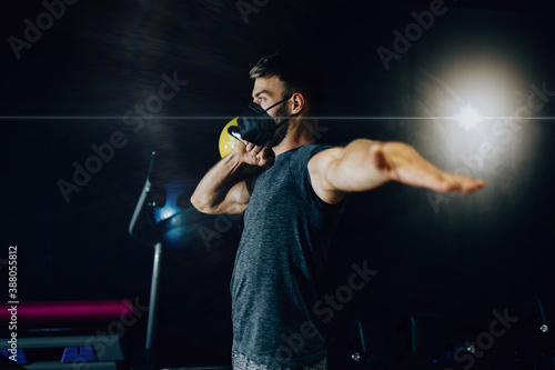 Fototapeta Naklejka Na Ścianę i Meble -  Good looking handsome male athlete with protective face mask exercising in modern fitness gym. Dark muddy light with strong shadows. Pandemic sports indoors concept.