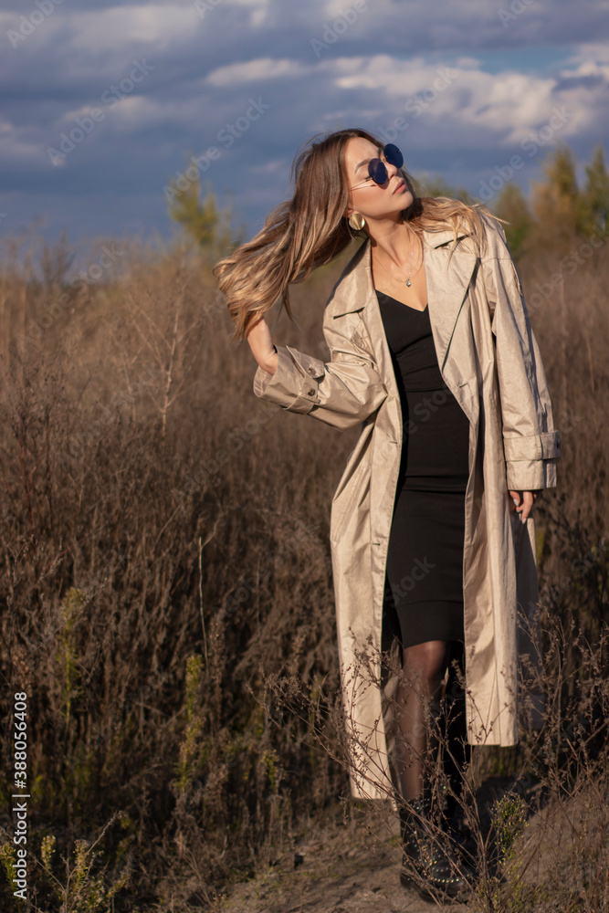 brunette in fashionable trench coat, black dress and sunglasses in the field. autumn, yellow leaves, blue sky. brunette in fashionable trench coat, black dress and sunglasses in the field. autumn, yellow leaves, blue sky.