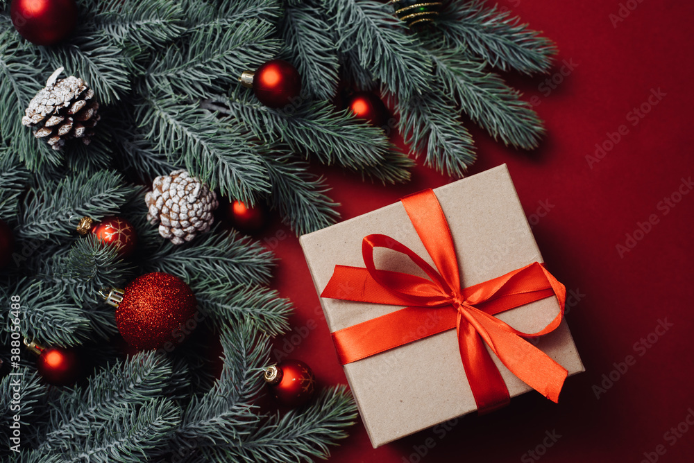 Craft gift with a red satin ribbon next to Christmas tree branches and balls, toys on a red background Craft gift with a red satin ribbon next to Christmas tree branches and balls, toys on a red background