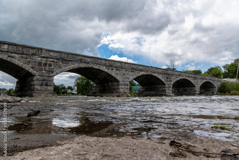 The historic Pakenham five-arch stone bridge in Pakenham, Ontario reflects in the river beneath it on a mostly cloudy day.