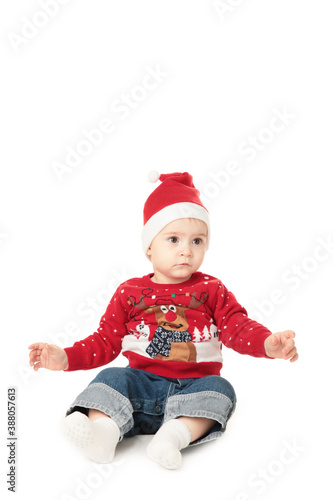 Adorable child is sitting on floor, wearing red Christmas hat, isolated over white