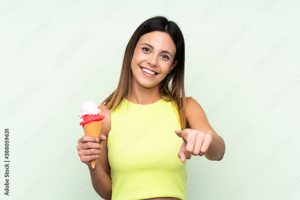 Brunette woman holding a cornet ice cream over isolated green background points finger at you with a confident expression Brunette woman holding a cornet ice cream over isolated green background points finger at you with a confident expression
