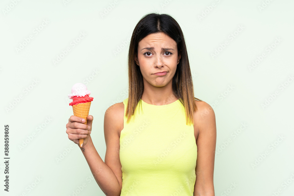 Brunette woman holding a cornet ice cream over isolated green background with sad expression Brunette woman holding a cornet ice cream over isolated green background with sad expression