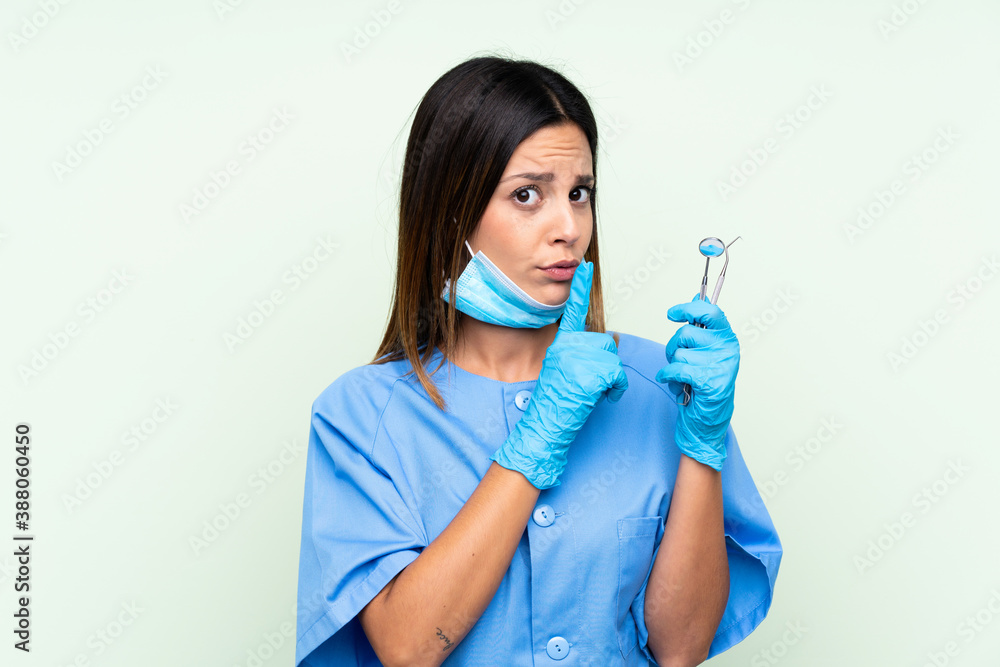 Woman dentist holding tools over isolated green background doing silence gesture Woman dentist holding tools over isolated green background doing silence gesture