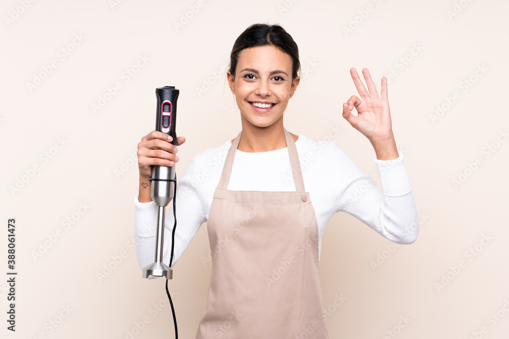 Woman using hand blender over isolated background showing ok sign with fingers Woman using hand blender over isolated background showing ok sign with fingers