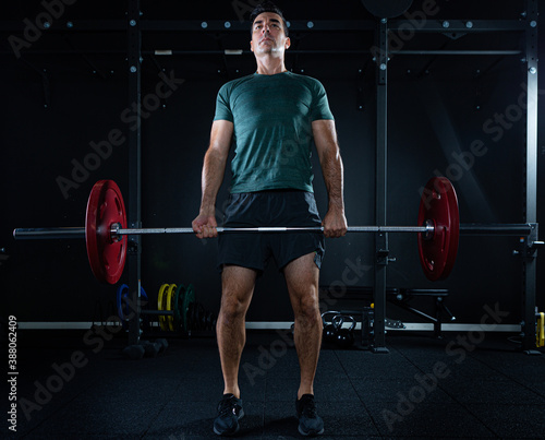 Tall dark man with green t shirt lifting barbell with red weights on a dark gym.