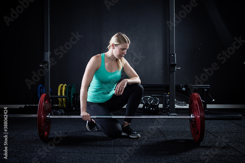 Blonde girl in green tank top in a dark gym resting a side barbell with red weights.