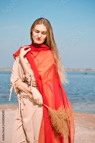 Young beautiful caucasian blond woman in light boho dress and red thin scarf, stays on beach by water under sunshine