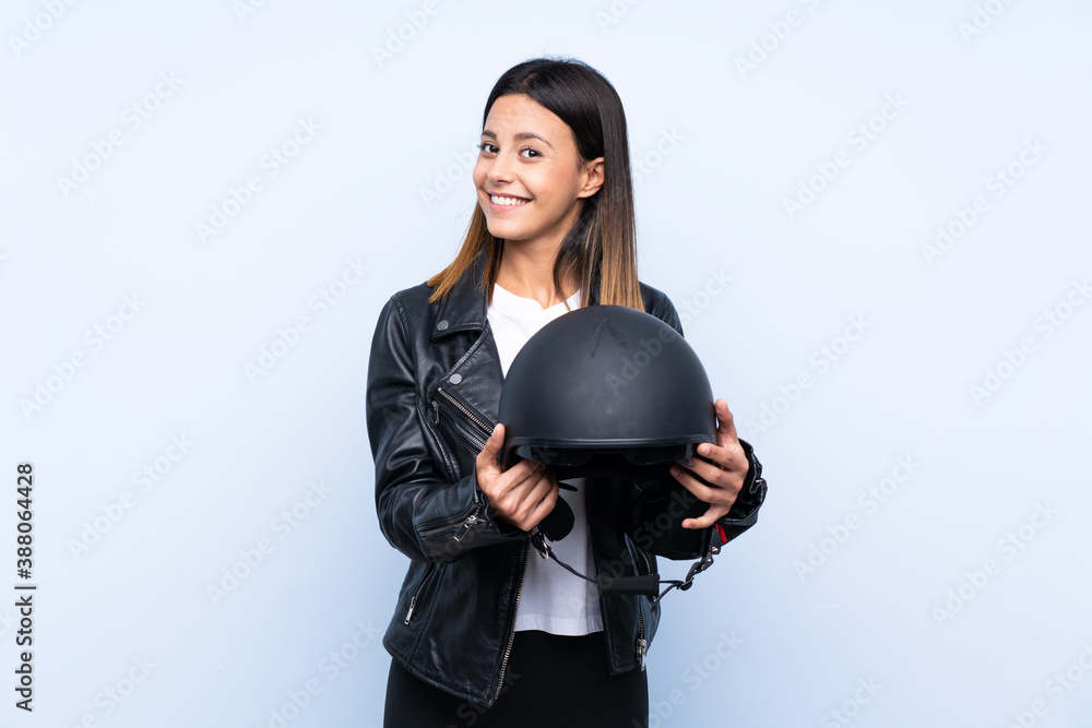 Young brunette woman holding a motorcycle helmet over isolated blue background with happy expression Young brunette woman holding a motorcycle helmet over isolated blue background with happy expression