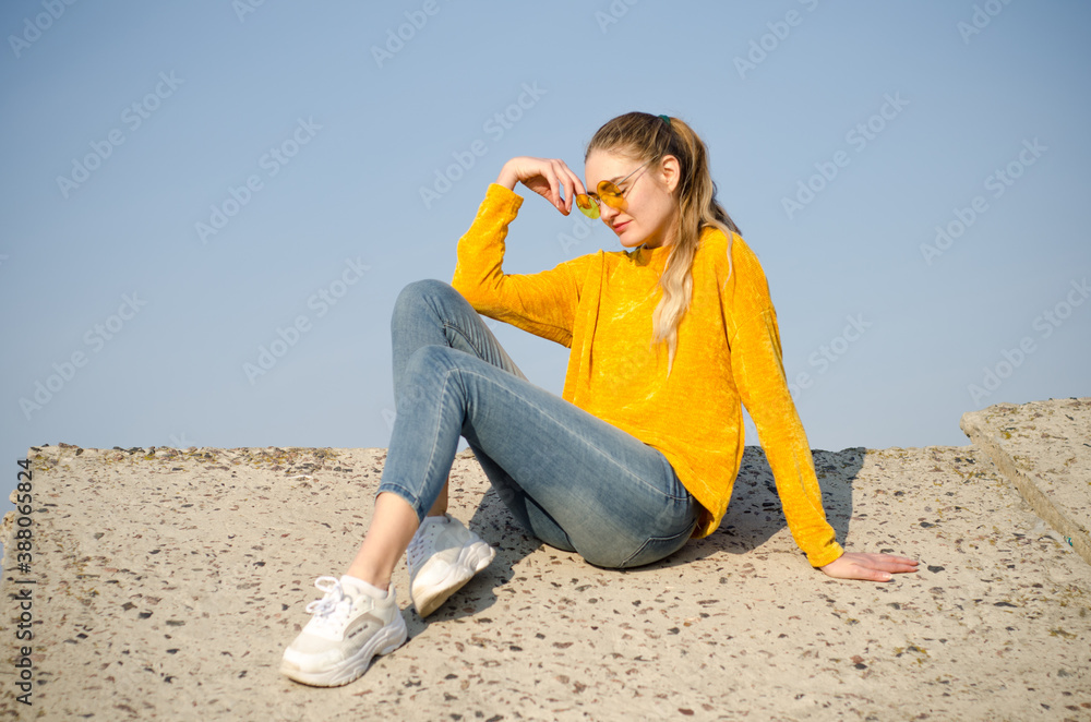 Beautiful blonde girl in yellow sweater and jeans, sneakers sits on concrete surface against blue sky in yellow glasses