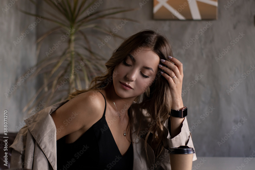 Fototapeta premium beautiful attractive slim brunette in trench coat in a grey loft room on leather couch with a paper cup. urban lifestyle concept. 