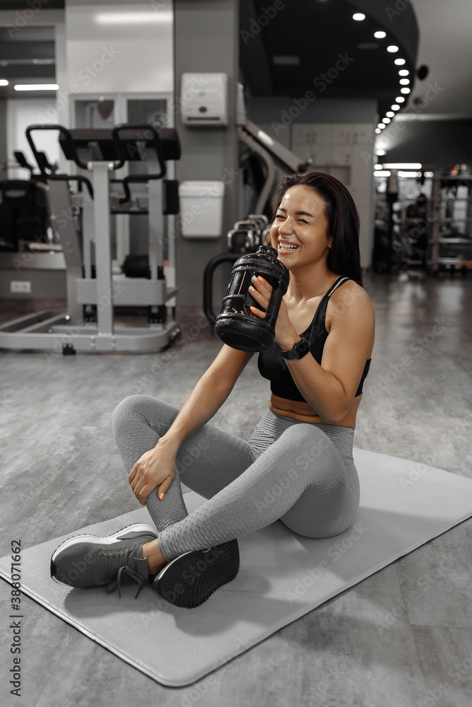 Smiling attractive sporty girl sitting on carpet, floor, having break, pause, drinking water in the gym. Health care and fitness concept Smiling attractive sporty girl sitting on carpet, floor, having break, pause, drinking water in the gym. Health care and fitness concept
